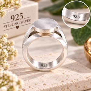 May include: A sterling silver ring featuring a flat circular top and a wide band. The interior of the band is stamped with the text 925. A circular inset shows a close-up of the inner band with the 925 hallmark clearly visible on the metal surface.