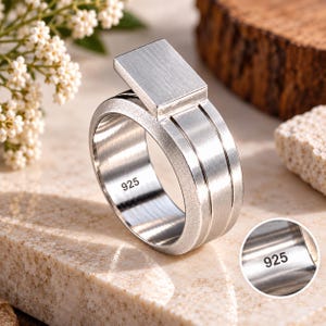 Könnte beinhalten: Ein silberner Ring mit gebürsteter Metalloberfläche und einer rechteckigen Kopfplatte. Die Innenseite des Rings ist deutlich mit dem Stempel 925 versehen, was auf Sterlingsilber hinweist. Das Design umfasst zwei parallele Rillen am äußeren Ring.