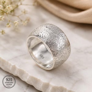 May include: A wide silver band ring featuring an intricate, raised floral pattern across its exterior surface. The interior of the band is smooth and clearly stamped with the hallmark 925, indicating sterling silver composition.