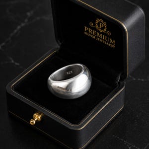 May include: A shiny, sterling silver ring with a rounded, domed shape, displayed in a black jewellery box. The ring is marked with "925" and rests on a black velvet interior. The box is embossed with "PREMIUM SILVER JEWELLERY" in gold lettering.