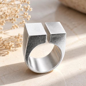 May include: A silver-coloured ring featuring a wide, open-top design with two distinct, flat-topped square blocks separated by a gap. The metal surface has a textured, matte finish on the exterior sides and a smooth, polished interior band.