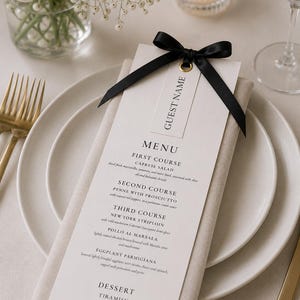 May include: A formal place setting with a menu card. The menu card is white with black text and a black ribbon. The menu lists courses such as Caprese Salad and New York Striploin. A gold fork and knife are visible.