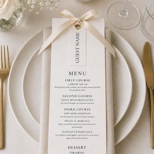 May include: A formal table setting with a menu card. The menu card is cream-colored with the words "GUEST NAME" and "MENU" printed on it. The menu lists courses like Caprese Salad and Tiramisu. The setting includes a plate, silverware, and a candle.