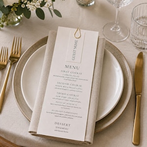 May include: Elegant table setting with a white plate, gold cutlery, and a folded linen napkin. A menu card with "GUEST NAME" and a detailed menu is held by a gold paper clip. A candle and flowers add to the ambiance.