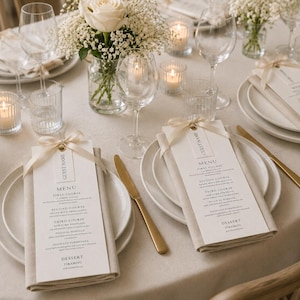May include: Elegant table setting with white plates, gold cutlery, and menu cards. The table is decorated with white flowers, candles, and wine glasses. Each menu card is tied with a ribbon and includes a guest name tag.
