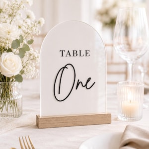 May include: An arched white acrylic table sign featuring the black printed text TABLE and the black script text One, supported by a rectangular light wood base. The sign is positioned upright, displaying a clear, smooth surface and clean edges.