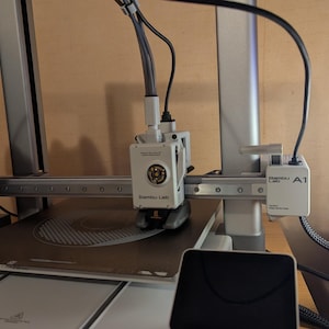 Può includere: Stampante 3D argento e nera con la scritta "Bambu Lab A1". Presenta un piano riscaldato e una testina di stampa che si muove lungo un asse orizzontale. Un piccolo schermo nero è in primo piano.
