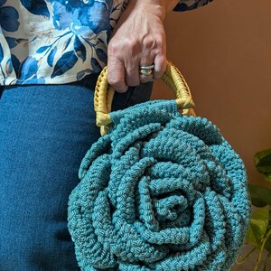 May include: Teal macrame handbag with a circular, rose-like pattern. The bag has a woven handle with a light yellow accent. The bag is held by a person wearing a blue floral top and blue jeans.