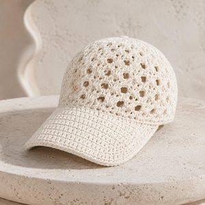 Puede incluir: Una gorra de béisbol de color crema hecha de hilo de ganchillo. La corona presenta un patrón de diamantes calado, mientras que la visera está construida con un punto de ganchillo denso y sólido. La gorra tiene una estructura redondeada.