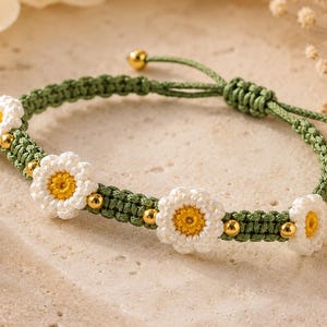 May include: A bracelet featuring a green woven cord base with four white and yellow crocheted flower motifs. The design includes small, polished gold-toned spherical beads interspersed between the flowers and at the ends of the adjustable cord.