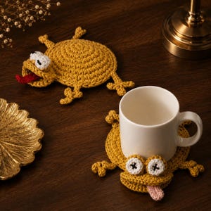 Peut inclure: Deux dessous de verre en forme de grenouille, faits à la main, en crochet jaune, avec des yeux blancs et noirs et des langues rouges et roses. Un dessous de verre avec une tasse blanche dessus. Sur une surface en bois brun foncé.