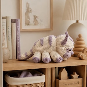 Puede incluir: Un juguete de peluche tejido a mano con un cuerpo de color crema con rayas de color púrpura claro, cuernos curvos y ojos grandes y oscuros. La criatura tiene seis patas, una cola larga a rayas y una cabeza redondeada con puntos visibles.