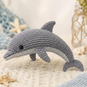 May include: A gray and light gray crocheted dolphin toy with a smooth, textured yarn surface, a prominent dorsal fin, two side flippers, a tail fluke, and a small black bead eye, crafted in a realistic shape with a curved body and pointed snout.