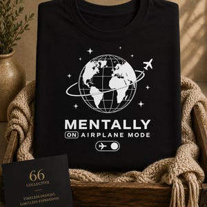May include: Black t-shirt featuring a white globe, airplane, and stars graphic. The text says "MENTALLY ON AIRPLANE MODE." A small card in the lower left corner reads "66 COLLECTIVE" and "TIMELESS DESIGNS. LIMITLESS EXPRESSION."