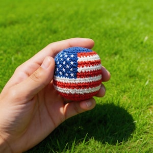 May include: A crocheted ball featuring the American flag design. The ball has a blue section with white stars, and red and white stripes. The ball is held against a backdrop of green grass.