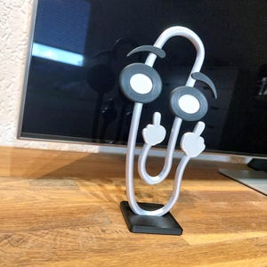 Op de afbeelding: Een grillige, 3D-geprinte sculptuur die lijkt op een paperclip met een gezicht. De figuur heeft grote, ronde ogen met witte centra, gebogen wenkbrauwen en twee vingerachtige aanhangsels. Het is wit met zwarte en grijze accenten en staat op een zwarte basis.