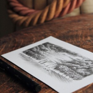 Puede incluir: Un bol&iacute;grafo negro se encuentra sobre una mesa de madera junto a un dibujo de una escena de bosque. El dibujo est&aacute; sobre papel blanco y muestra un bosque con &aacute;rboles y un cielo nublado.