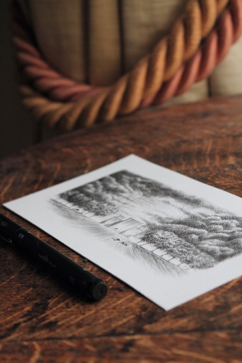 Puede incluir: Un bol&iacute;grafo negro se encuentra sobre una mesa de madera junto a un dibujo de una escena de bosque. El dibujo est&aacute; sobre papel blanco y muestra un bosque con &aacute;rboles y un cielo nublado.