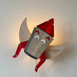 Könnte beinhalten: Eine Holzraketenlampe mit roter Nase und Beinen, vor einem Halbmond. Die Rakete hat ein lächelndes Gesicht und ist an einer Wand befestigt. Die Lampe ist für ein Kinderzimmer konzipiert. Der Mond hat den Namen "Jonathan" und das Datum "01-08-2020".