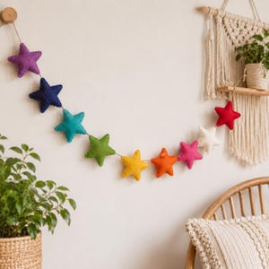 May include: A decorative garland featuring ten plush, stuffed stars in a vibrant rainbow spectrum of colors including purple, blue, teal, green, yellow, orange, pink, white, and red, all strung together on a thin, natural-colored twine cord.