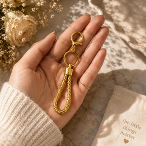 May include: A gold-coloured keychain with a braided leather-like handle and a clasp. The keychain is held in a hand, with a small bag in the background that reads "the little things matter".