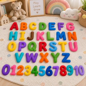 May include: A set of plush, multi-colored felt letters from A to Z and numbers from 0 to 10 arranged on a white circular rug. Each character features a soft, stuffed construction with distinct colors including red, orange, yellow, green, blue, purple, pink, and brown.
