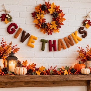 May include: A felt letter garland spelling GIVE THANKS in autumn colors, hung below a wreath of maple leaves, pinecones, and berries. The display includes a lantern with a lit candle, pumpkins, and foliage on a wooden mantelpiece.