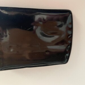 May include: A rectangular, black, patent leather wallet with rounded corners. The wallet has a glossy finish and reflects light, showing slight imperfections on the surface. The edges are neatly stitched.