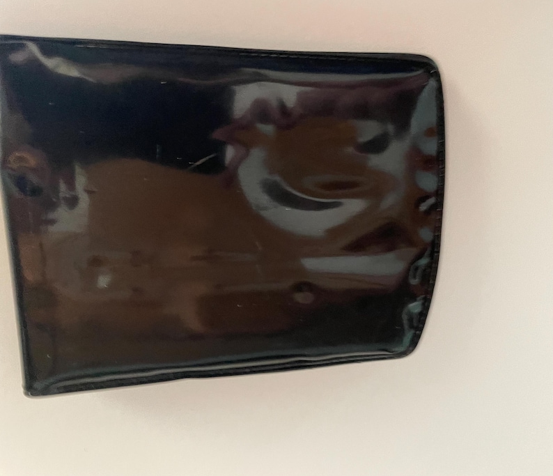 May include: A rectangular, black, patent leather wallet with rounded corners. The wallet has a glossy finish and reflects light, showing slight imperfections on the surface. The edges are neatly stitched.