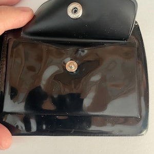 May include: Black vinyl wallet with a snap closure. The wallet is open, revealing a clear plastic pocket and a small pocket. The snap is silver-colored. The wallet is likely designed to hold cards or cash.