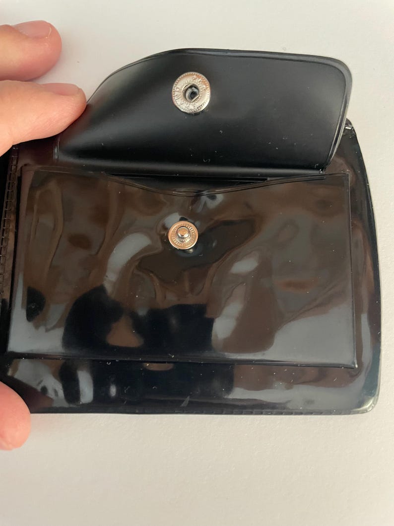 May include: Black vinyl wallet with a snap closure. The wallet is open, revealing a clear plastic pocket and a small pocket. The snap is silver-colored. The wallet is likely designed to hold cards or cash.