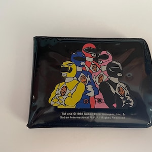 May include: A black wallet featuring a colorful graphic of the Power Rangers. The image shows the Rangers in their signature helmets and uniforms. The wallet has a glossy finish and includes text from 1993 Saban Entertainment.