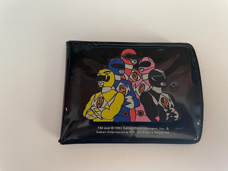 May include: A black wallet featuring a colorful graphic of the Power Rangers. The image shows the Rangers in their signature helmets and uniforms. The wallet has a glossy finish and includes text from 1993 Saban Entertainment.