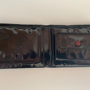 May include: Open black wallet with a glossy, patent leather finish. The wallet has a small pocket with a red button closure on the right side. The interior is visible, showing the open compartments.