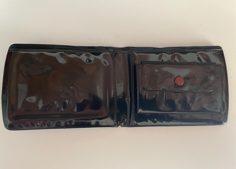 May include: Open black wallet with a glossy, patent leather finish. The wallet has a small pocket with a red button closure on the right side. The interior is visible, showing the open compartments.