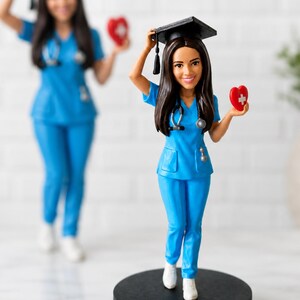 Może przedstawiać: Personalizowana figurka z kiwającą głową przedstawiająca pracownika służby zdrowia w niebieskim stroju, białych butach i czarnym birecie. Figurka trzyma czerwone serce z białym krzyżem i ma stetoskop zawieszony na szyi.