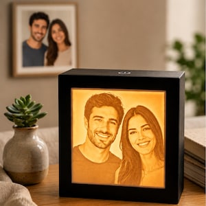 May include: A square, black-framed light box featuring a warm, glowing, monochromatic lithophane-style portrait of a smiling man and woman. The device has a power button on the top edge and emits a soft, amber-toned light from the front panel.