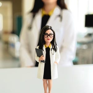 Op de afbeelding: Een doktersfiguur met lang donker haar en een bril met zwart montuur, gekleed in een witte doktersjas over een zwarte jurk met een gele kraag. Het figuurtje houdt een donkergrijs klembord vast, draagt een stethoscoop en staat op een zwarte voet.