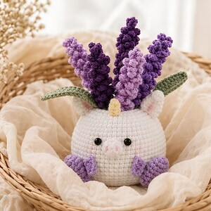 Könnte beinhalten: Ein weißes, gehäkeltes Einhorn-Stofftier mit schwarzen Augen, einem kleinen gelben Horn und violetten Hufen. Das Einhorn hat mehrere hohe, texturierte, violette und hellviolette gehäkelte Lavendelzweige auf dem Kopf, akzentuiert durch zwei grüne Blätter.