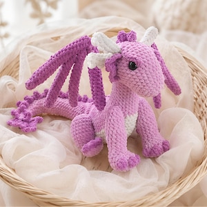 May include: A handmade crochet dragon in shades of purple, with white accents on the horns and chest. The dragon has large wings and a long tail, and is sitting in a woven basket lined with white fabric. The dragon is a whimsical, soft toy.