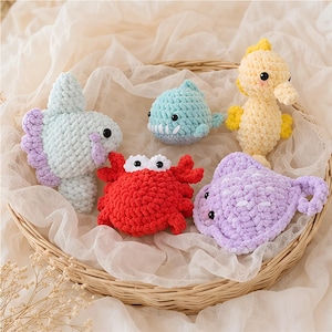 May include: A collection of handmade crochet sea creatures, including a light blue fish, a blue shark, a yellow seahorse, a red crab, and a purple fish. The toys are displayed in a woven basket, showcasing their soft textures and playful designs.