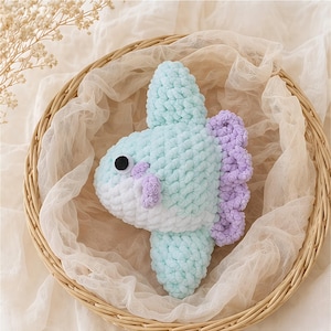 May include: A handmade crochet fish in a wicker basket. The fish features a light blue body, white belly, and lavender fins. It has a black eye and is made of soft, textured yarn. The basket is lined with a sheer, cream-colored fabric.