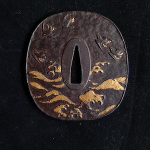 May include: A dark brown, oval-shaped metal sword guard, known as a tsuba, featuring a textured surface with gold-coloured accents depicting stylised waves at the bottom and birds in flight across the upper portion, with a central tapered opening.