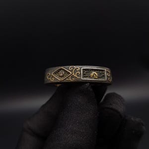 May include: A dark metal band ring featuring a textured surface with gold-colored inlay details. The design includes a diamond-shaped frame containing a small circular element and a rectangular frame with a curved gold-colored motif, accented by swirls.