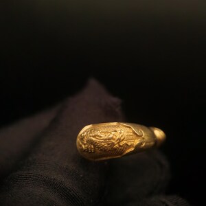 May include: A gold-colored, ornate ring with a detailed floral design. The ring has a rounded shape with a textured surface. The background is dark, emphasizing the ring's intricate details and golden hue.