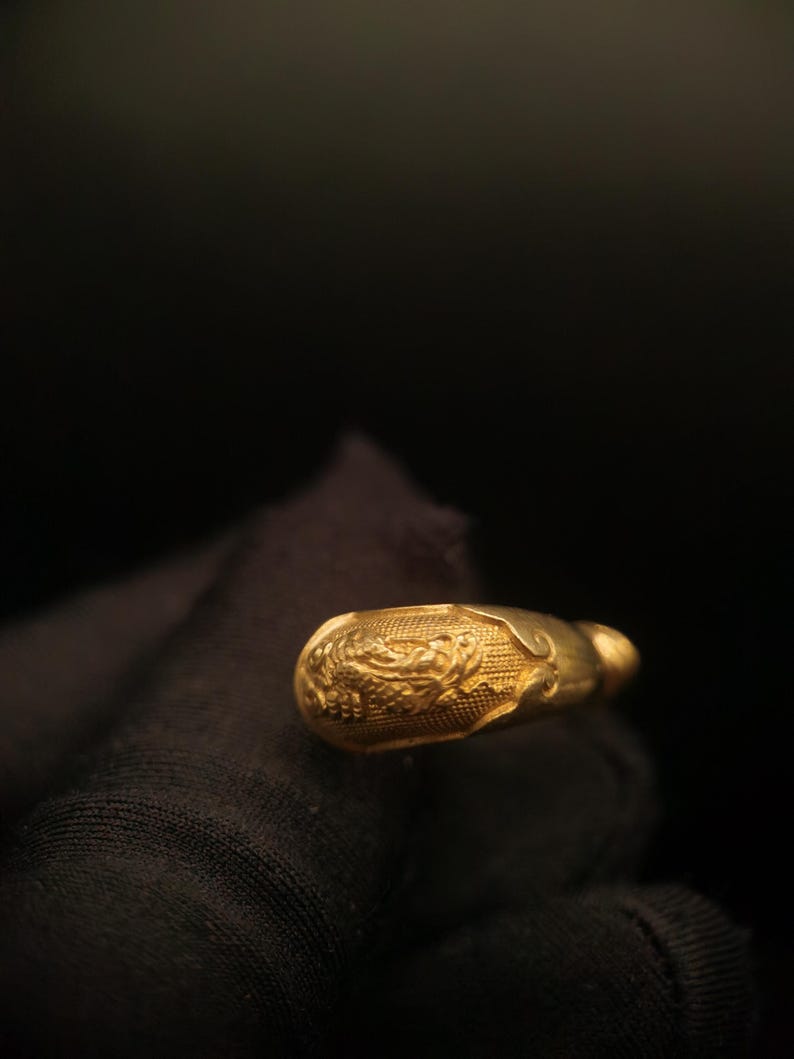 May include: A gold-colored, ornate ring with a detailed floral design. The ring has a rounded shape with a textured surface. The background is dark, emphasizing the ring's intricate details and golden hue.