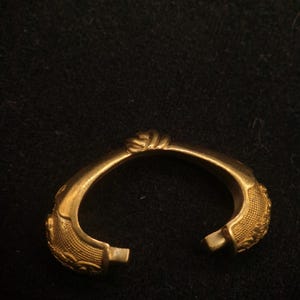 May include: A gold-colored, open-ended bracelet with ornate detailing. The bracelet features textured patterns and a twisted knot design at the top. The ends have small, rectangular clasps. The jewelry is set against a dark background.
