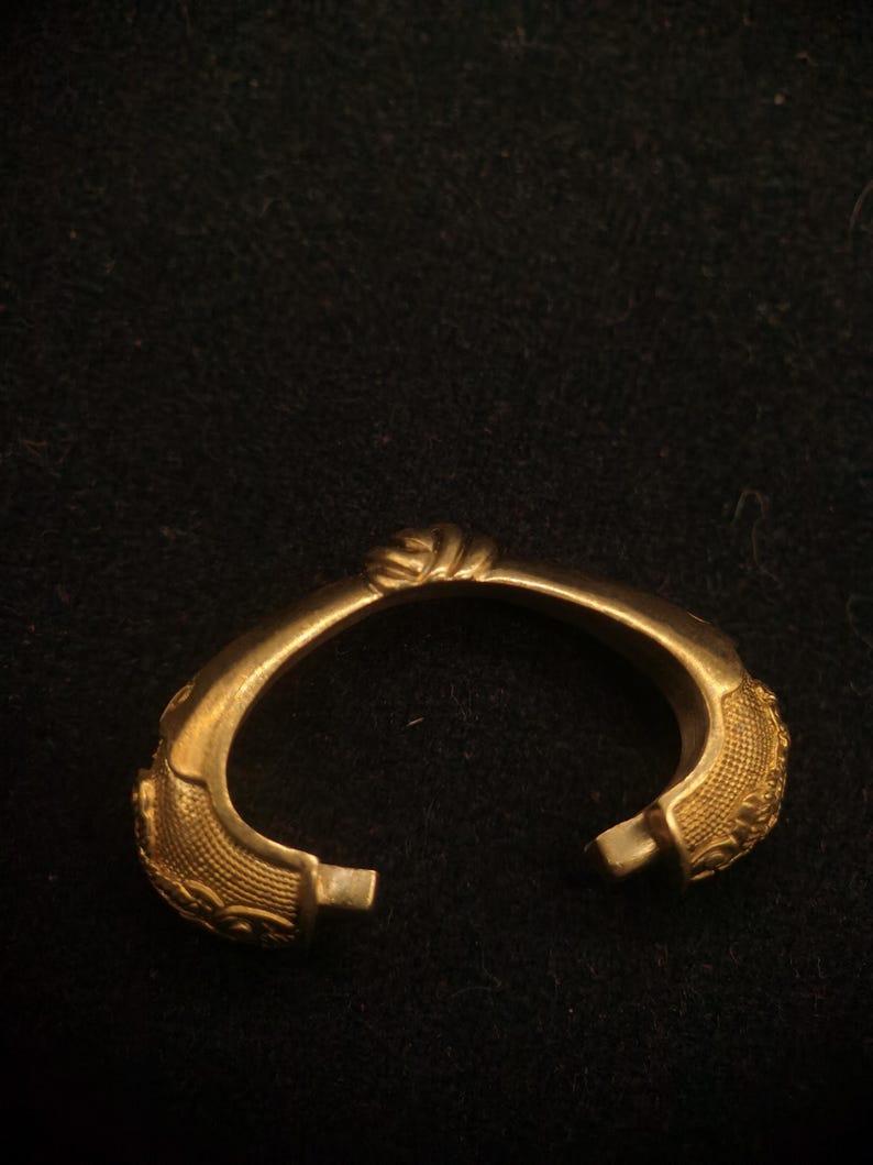May include: A gold-colored, open-ended bracelet with ornate detailing. The bracelet features textured patterns and a twisted knot design at the top. The ends have small, rectangular clasps. The jewelry is set against a dark background.