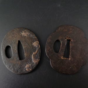 May include: Two dark brown, weathered metal sword guards, known as tsuba. The left guard is oval with a textured surface and floral relief, while the right guard features a four-lobed shape. Both contain central and side cutouts for blade fitting.