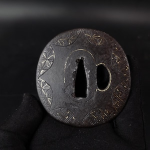 May include: A circular dark metal sword guard, known as a tsuba, featuring an etched geometric star pattern and a grid design along the outer edge. The centre contains a large tapered opening and a smaller adjacent oval hole for blade mounting.